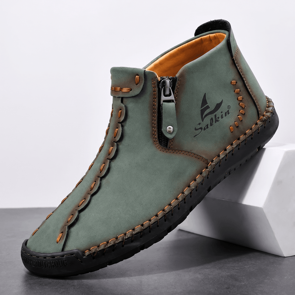 Single sage green suede Javier high-top shoe with contrast orange stitching and side zipper, angled on a white block against a studio backdrop
