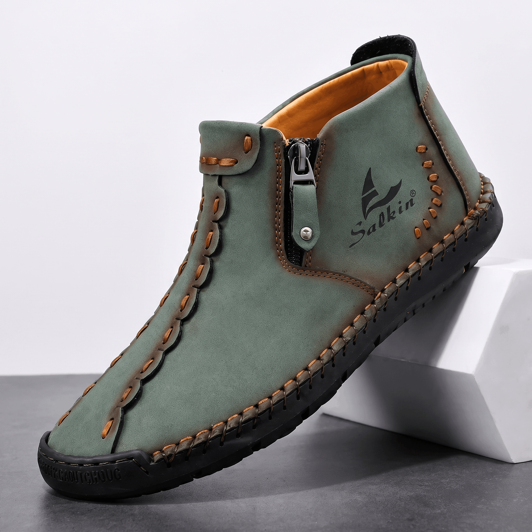 Single sage green suede Javier high-top shoe with contrast orange stitching and side zipper, angled on a white block against a studio backdrop