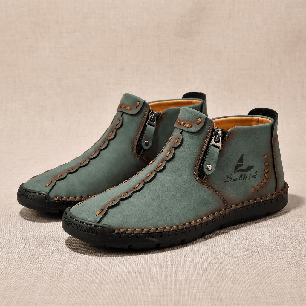 Pair of sage green suede Javier high-top shoes with orange stitching and zippers, resting on a beige fabric backdrop