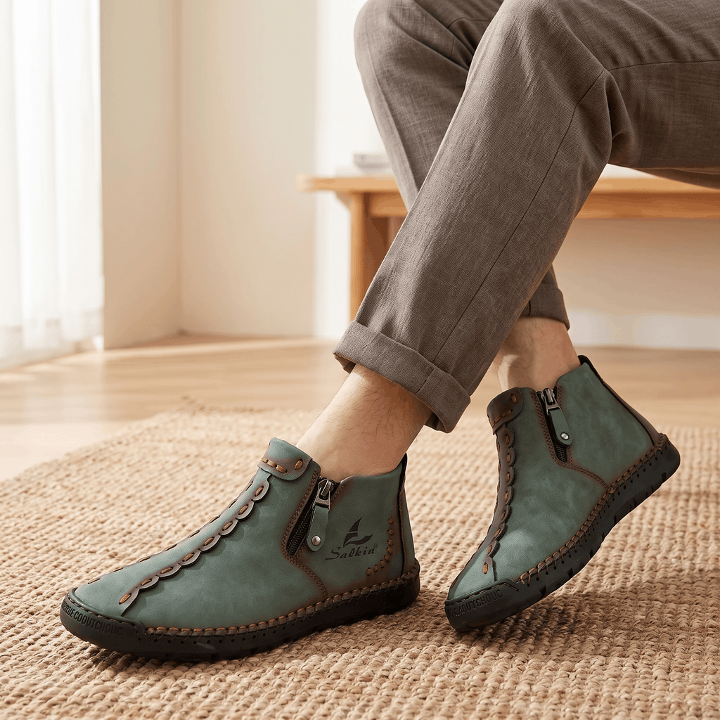 Close-up of crossed ankles wearing gray trousers and sage green suede Javier high-top zip shoes on a jute rug near a window