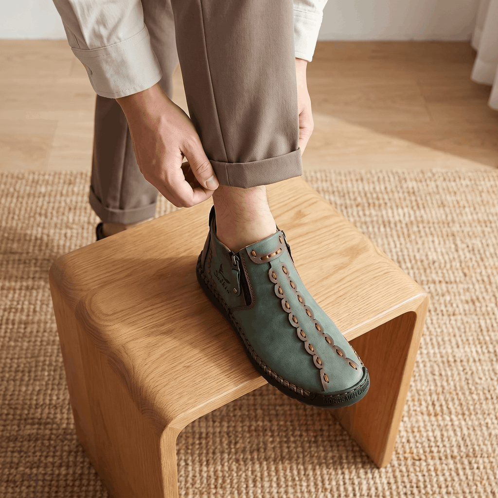 Man adjusting trouser cuff with foot propped on wooden stool, wearing sage green suede Javier high-top shoe over a jute rug