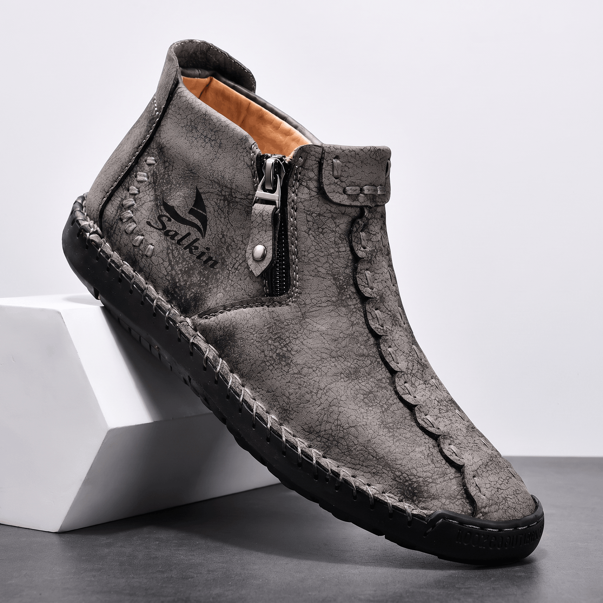 Single gray suede Javier high-top shoe with side zipper and hand-stitched trim, angled on a white block against a studio backdrop