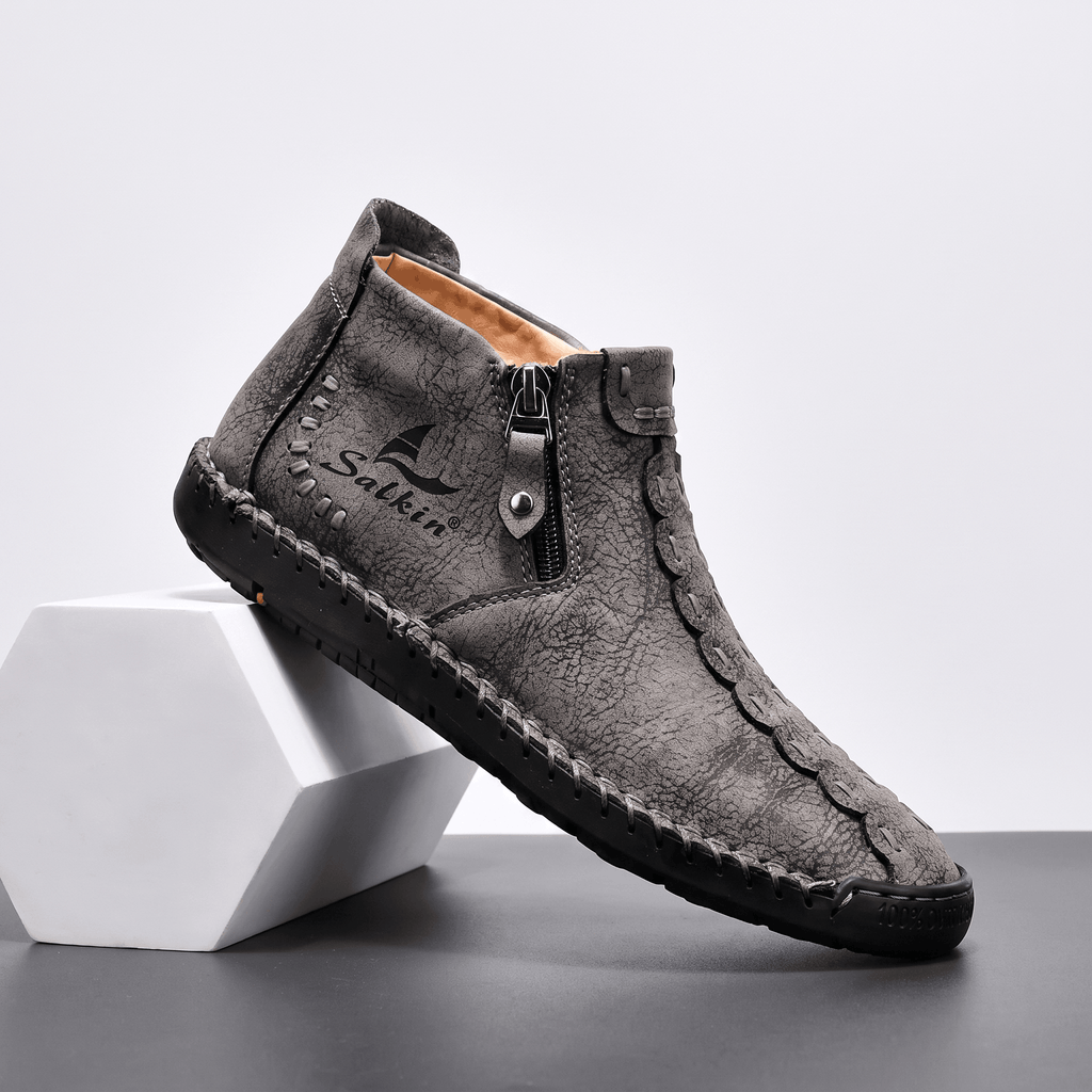 Single gray suede Javier high-top shoe propped against a hexagonal white block, showing side zipper and scalloped stitched panel