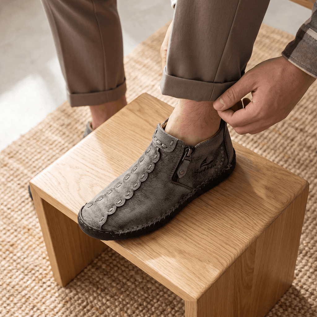 Man with foot resting on wooden stool adjusting sock, wearing gray suede Javier high-top shoe on a jute rug