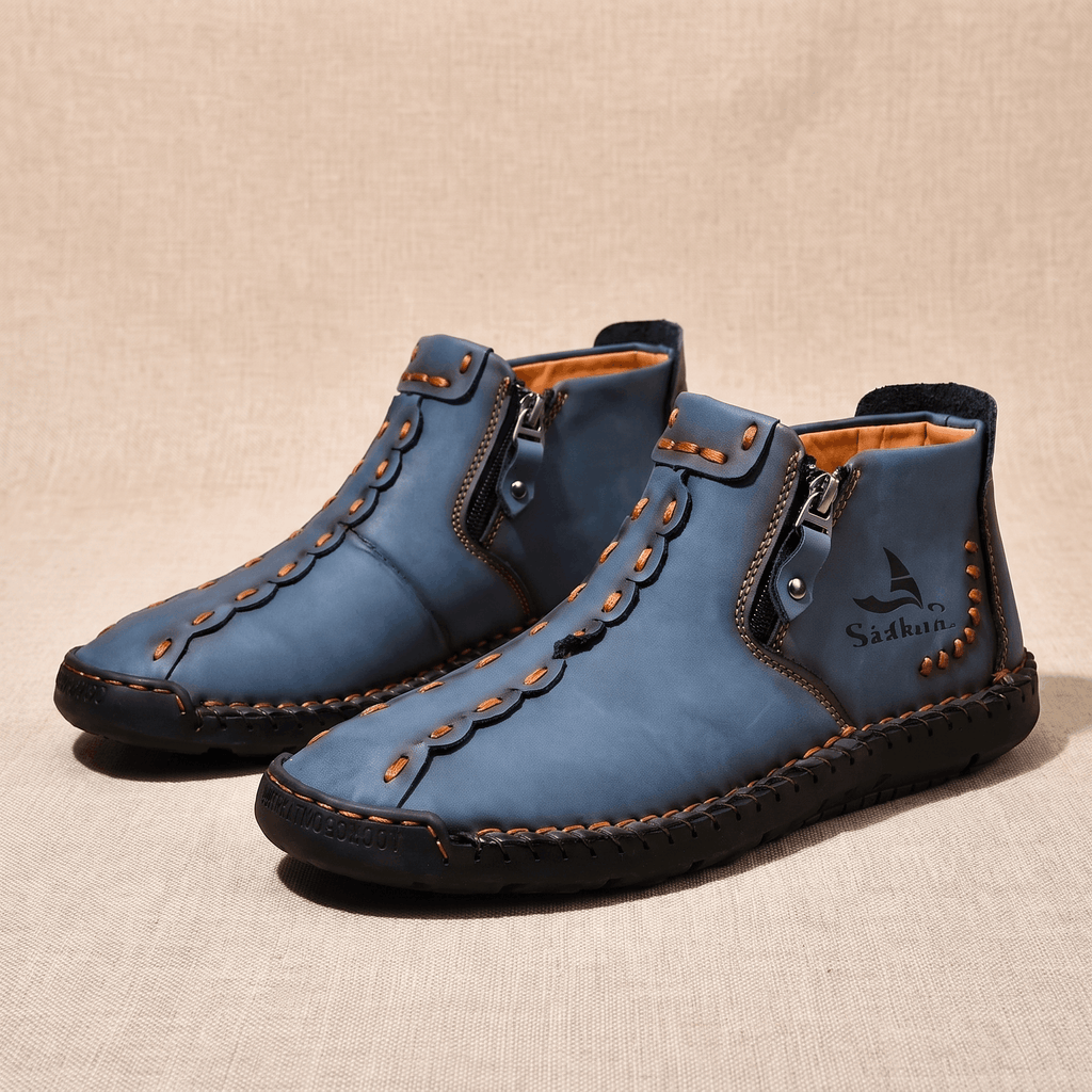 Pair of blue leather Javier high-top shoes with orange stitching and zippers, resting on a beige fabric backdrop