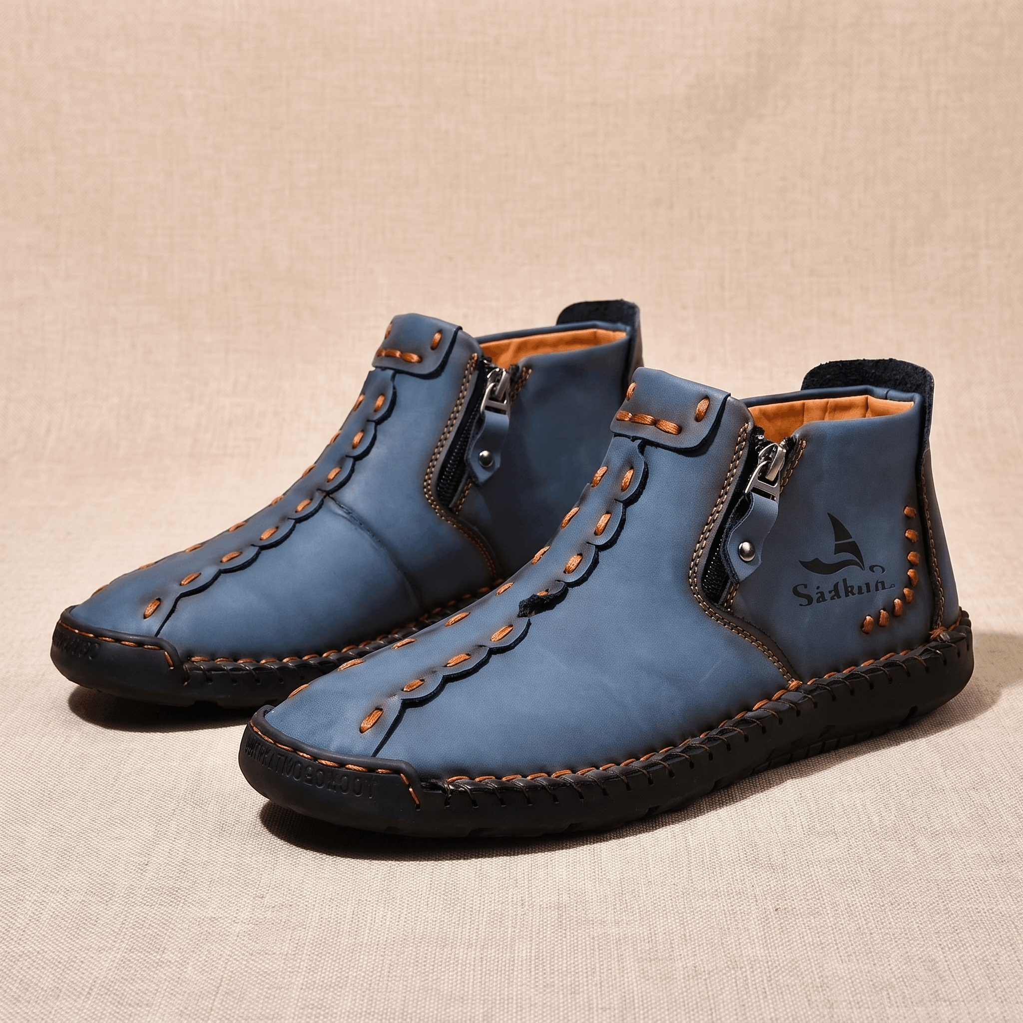 Pair of blue leather Javier high-top shoes with orange stitching and zippers, resting on a beige fabric backdrop