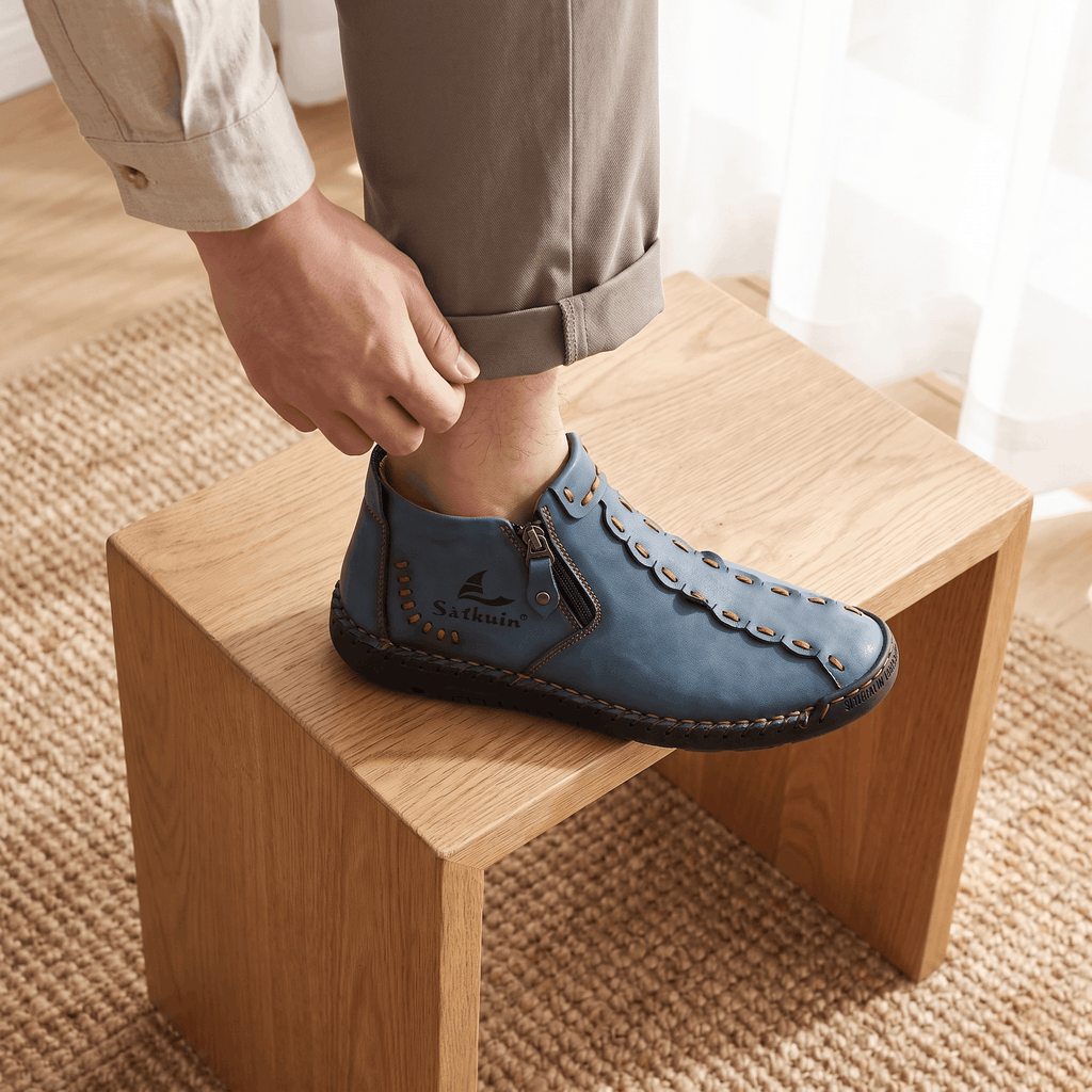 Man adjusting cuff with foot resting on wooden block wearing blue leather Javier high-top shoe with orange stitching, near a curtained window