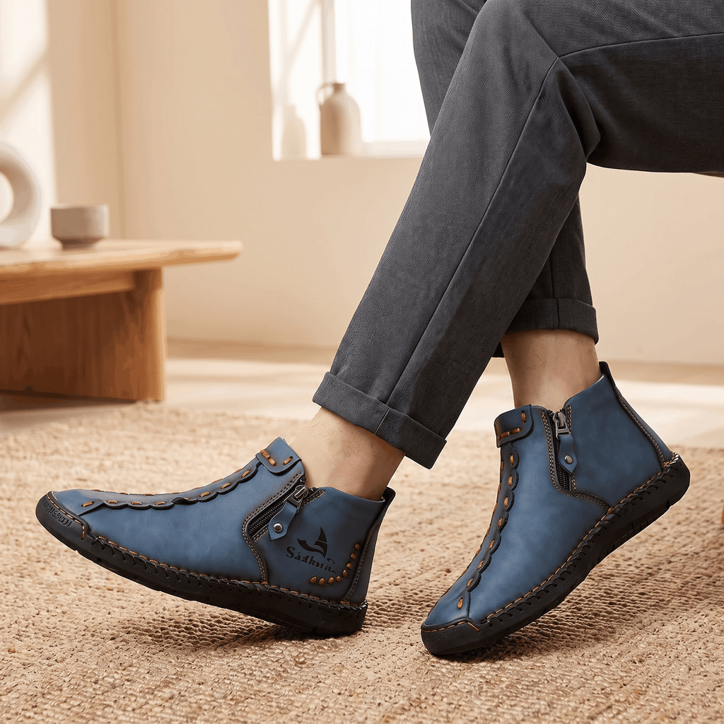 Close-up of crossed ankles wearing gray trousers and blue leather Javier high-top zip shoes on a jute rug near a low wooden table