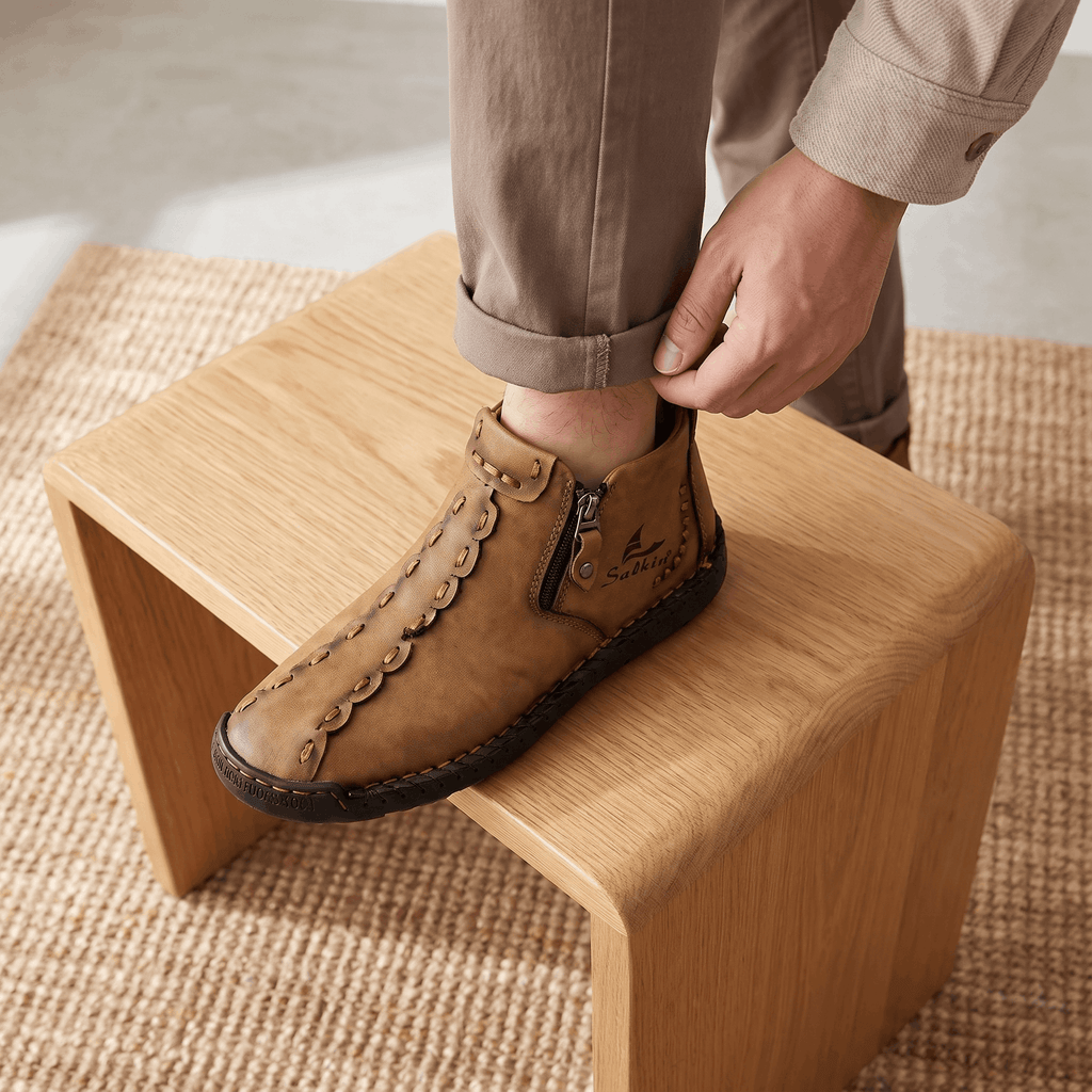 Man adjusting trouser cuff with foot resting on wooden stool wearing tan leather Javier high-top shoe over jute rug
