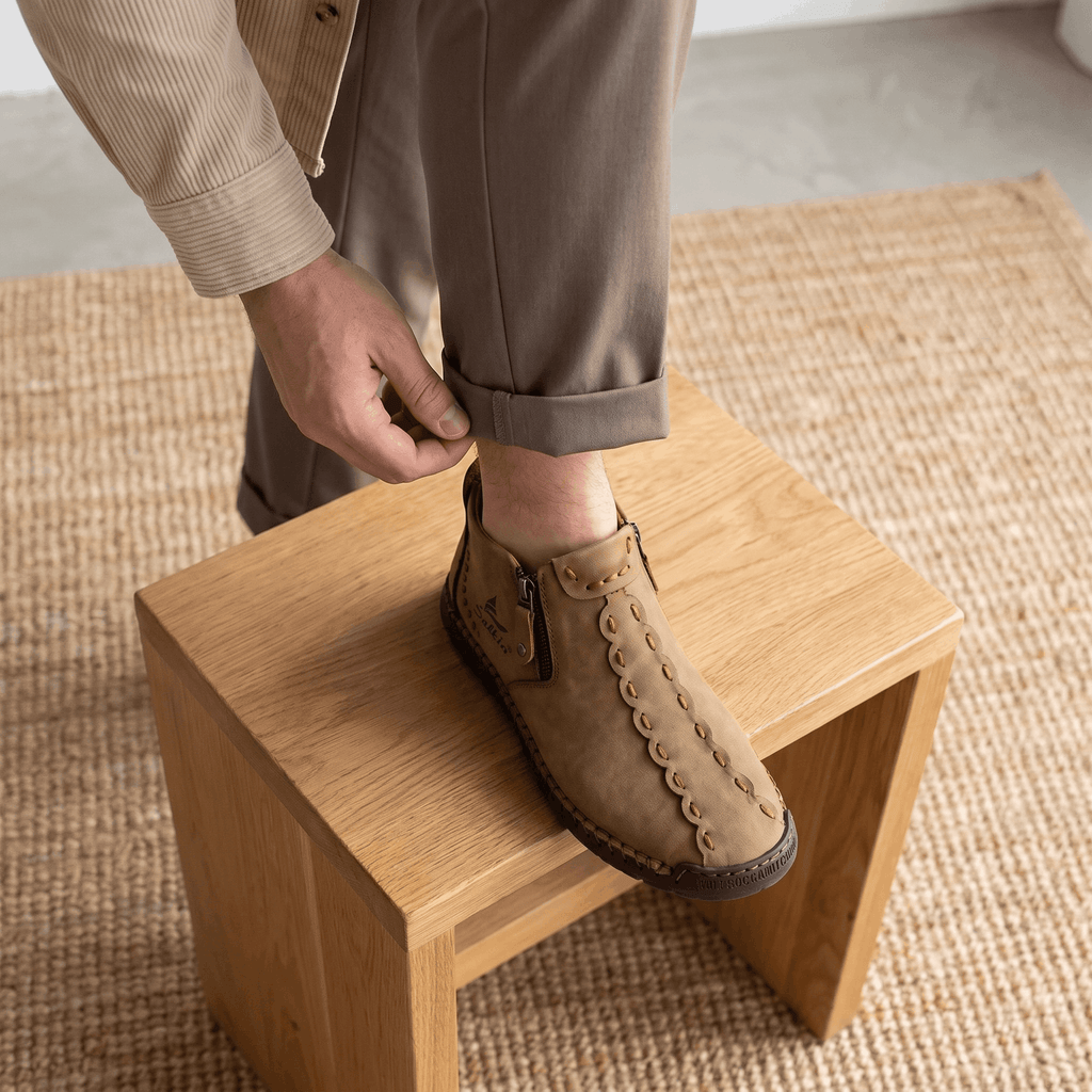 Man adjusting trouser cuff with foot resting on wooden stool wearing light brown leather Javier high-top shoe over jute rug