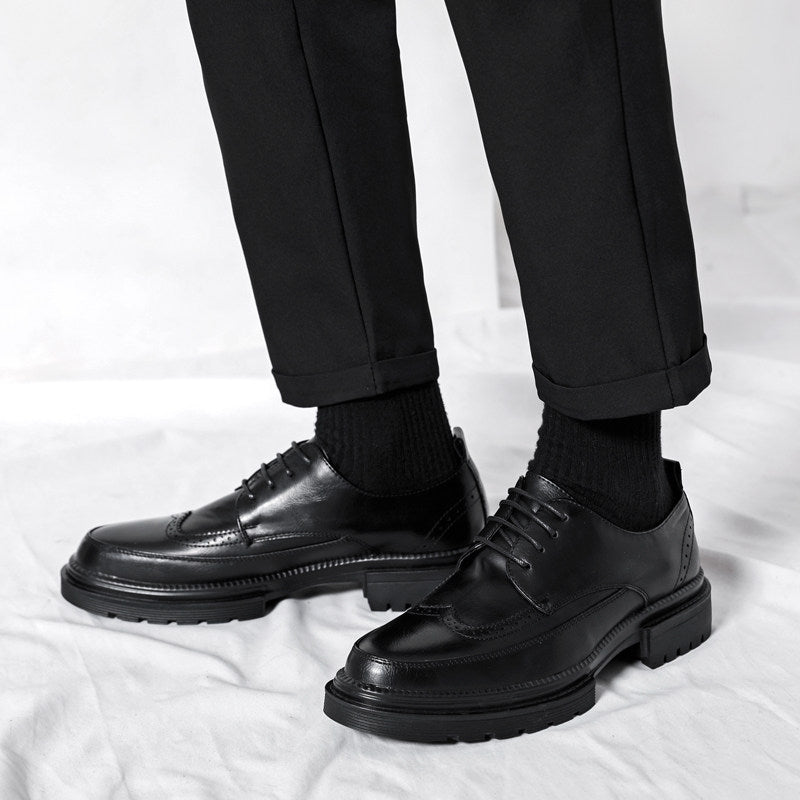 Man's legs in black tailored trousers and black socks wearing black leather Julian brogue dress shoes, standing on a white backdrop