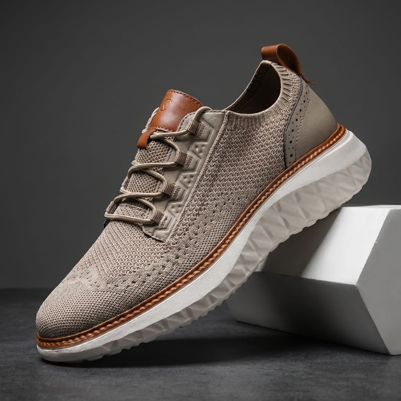 Khaki mesh sneaker with brown leather trim and white cushioned sole propped diagonally against a white geometric block