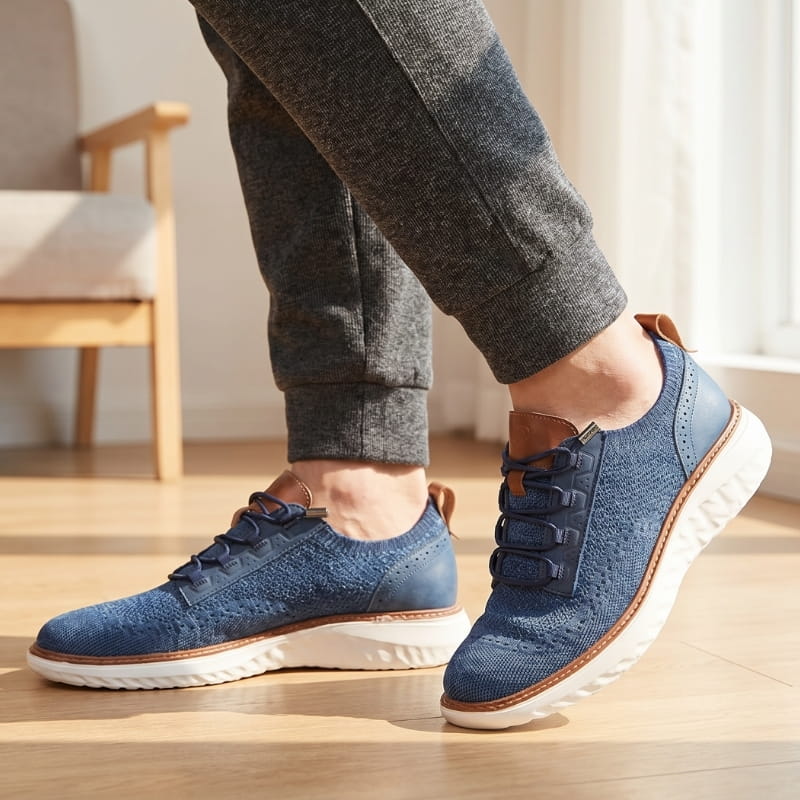 Person wearing navy blue mesh sneakers with brown trim and gray trousers, standing on a wood floor indoors