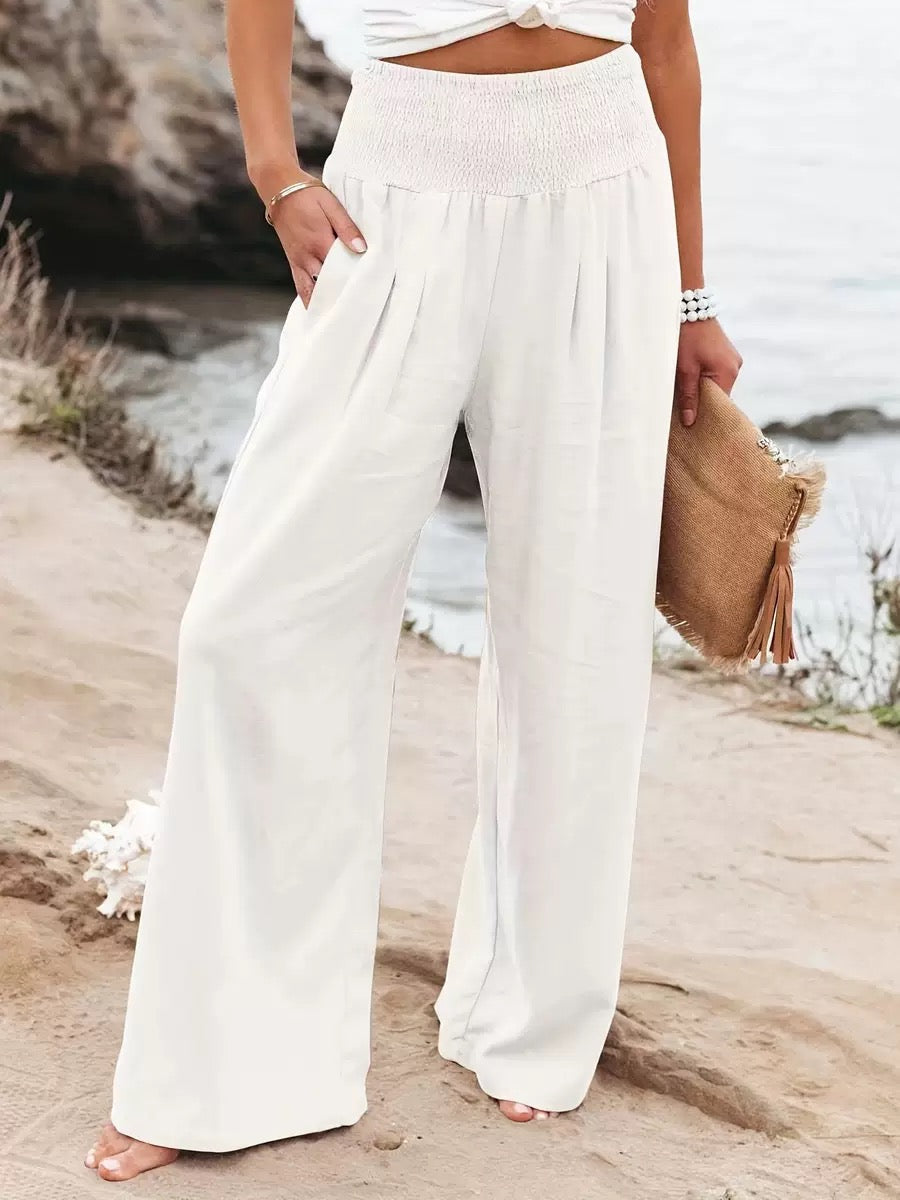 Women's white high-waisted wide-leg linen pants with a flowy silhouette, elastic waistband, and side pockets in a beach setting.