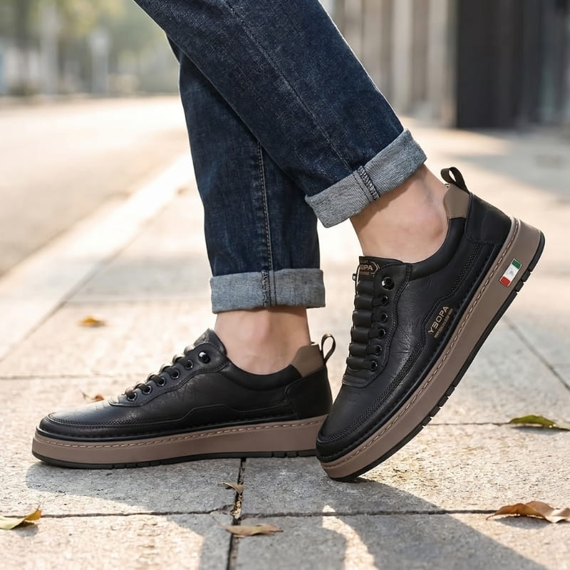 Person wearing black leather chunky sneakers with cuffed jeans, walking on a paved outdoor path with fallen leaves