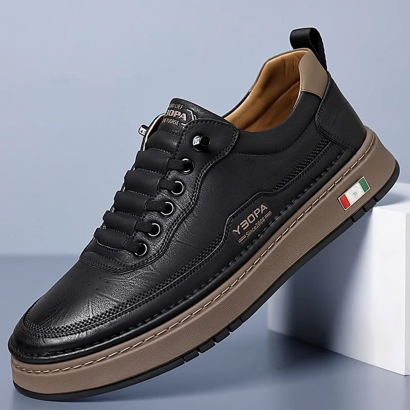 Black leather chunky sneaker with tan sole propped diagonally against a white block on a blue studio background