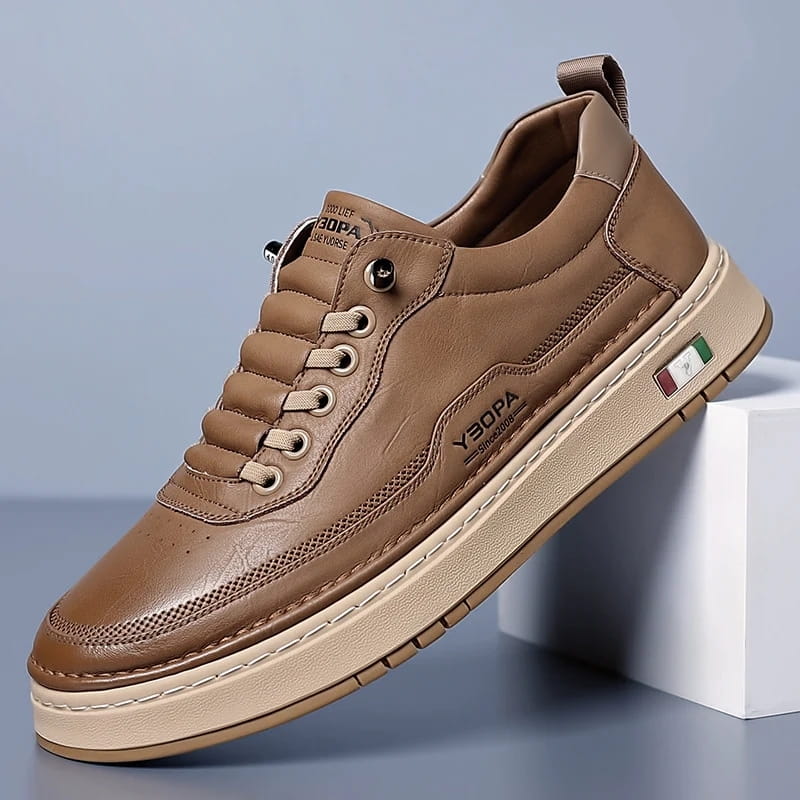 Brown leather chunky sneaker with tan sole and side patch detail propped diagonally against a white block on a blue studio background