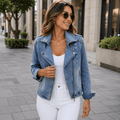 Woman wearing a light wash denim moto jacket with an asymmetrical zip closure, white top, white jeans, and sunglasses while walking in an urban setting.