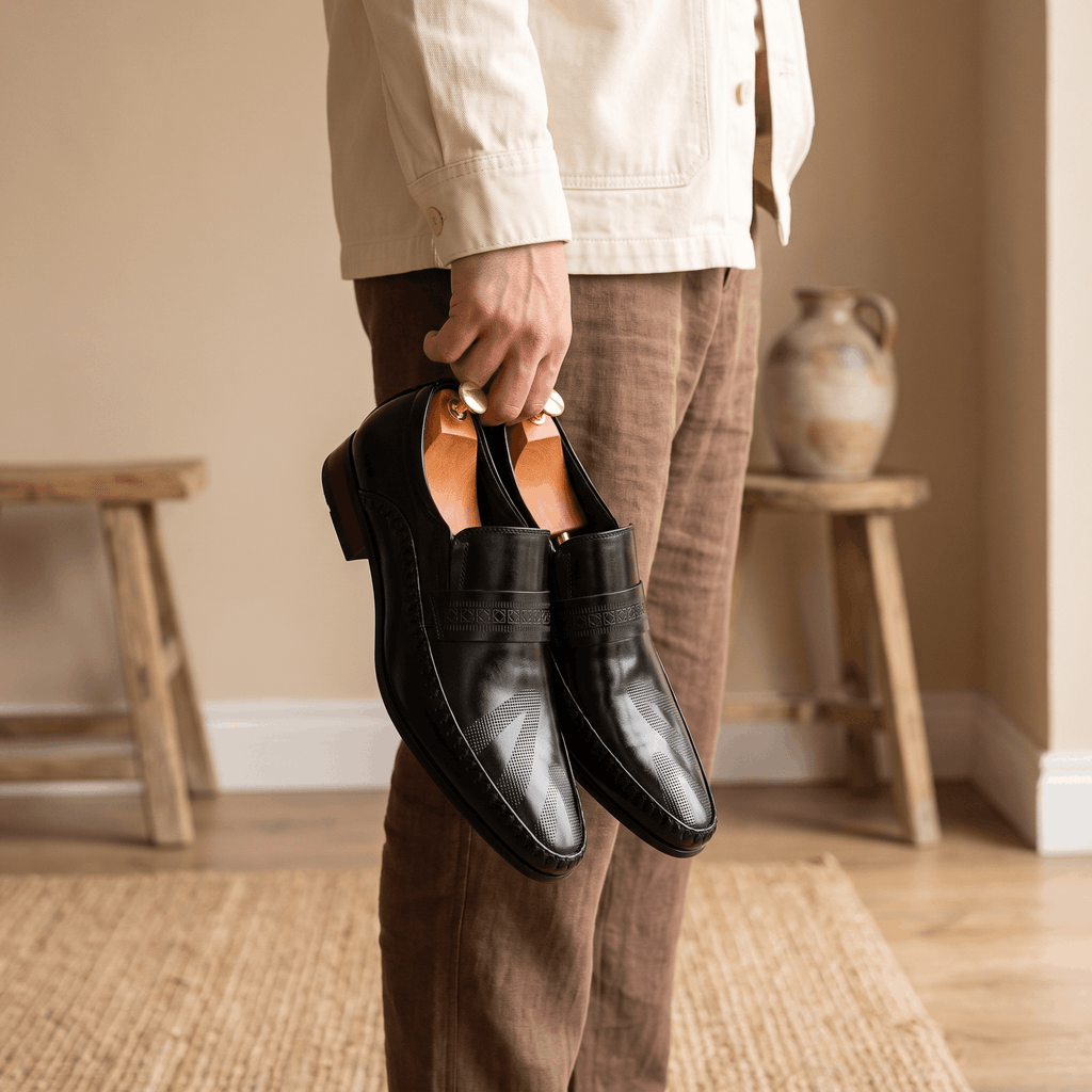 Man in cream shirt and brown trousers holding a pair of black patent leather Leonardo loafers by hand indoors