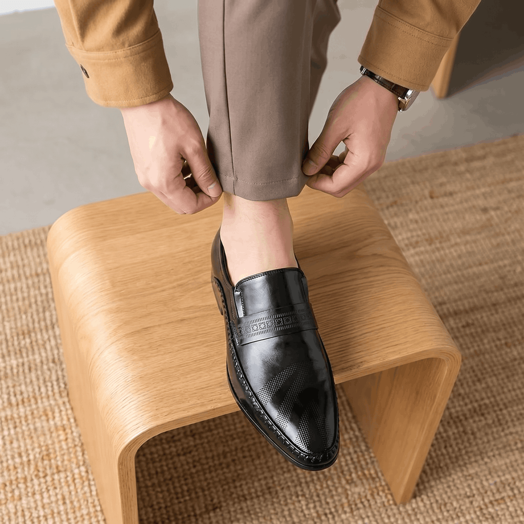 Man adjusting trouser cuff with foot resting on curved wooden stool wearing black patent leather Leonardo loafer over jute rug