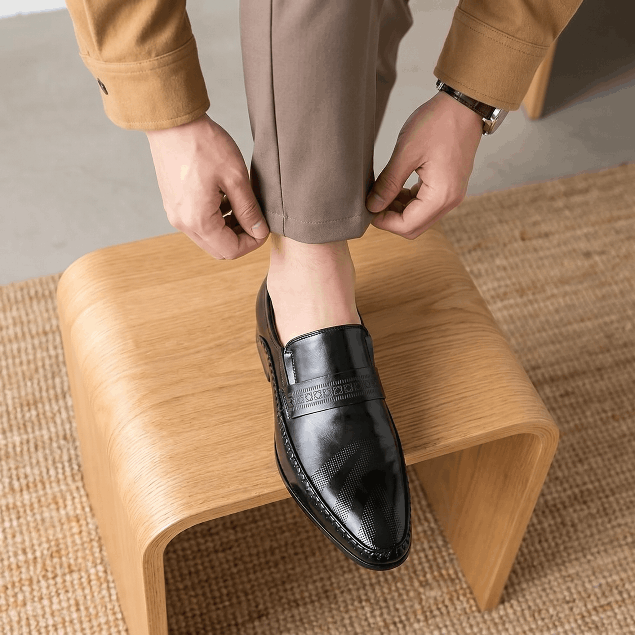 Man adjusting trouser cuff with foot resting on curved wooden stool wearing black patent leather Leonardo loafer over jute rug