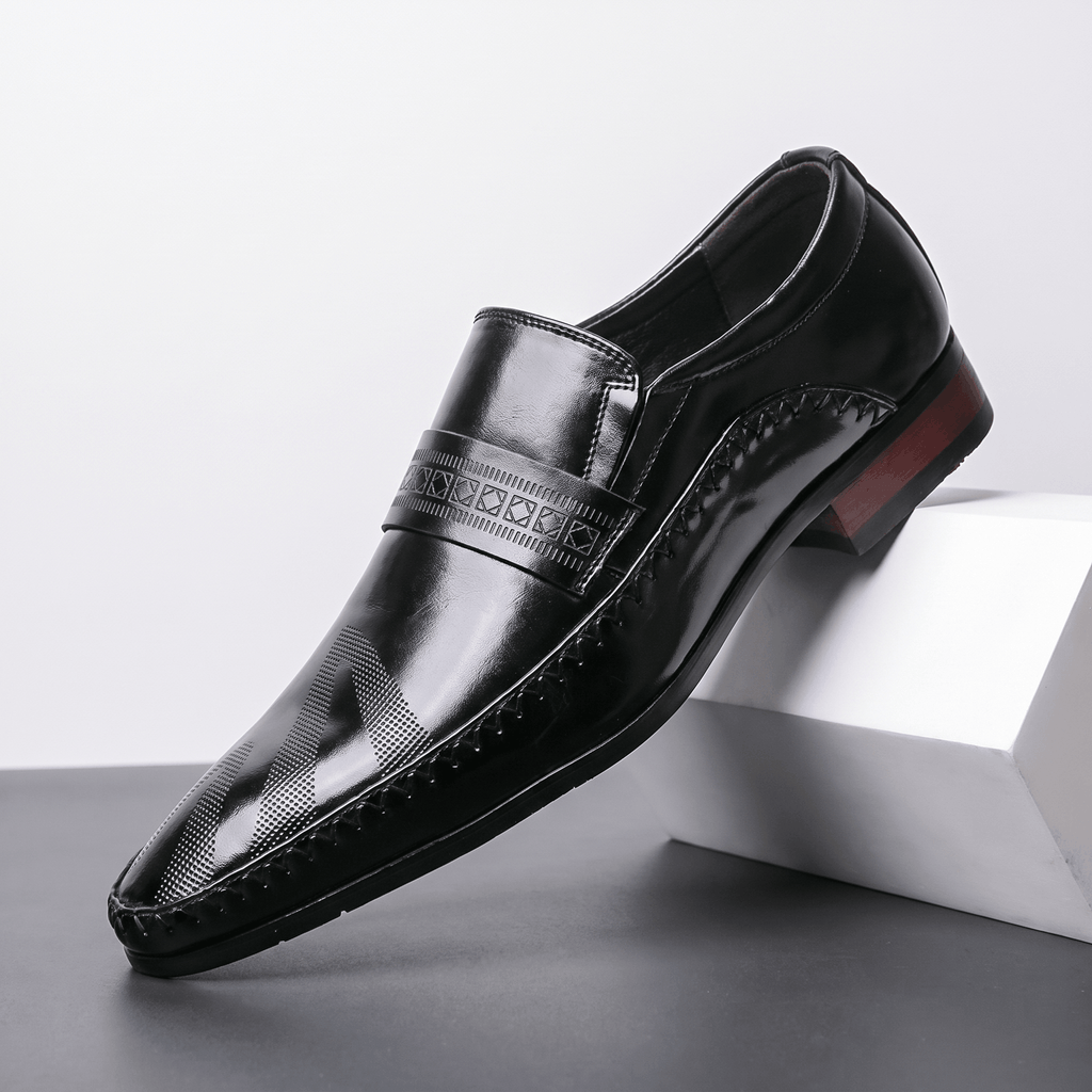 Single black patent leather Leonardo loafer with etched band and burgundy heel accent, propped against a white block in studio setting