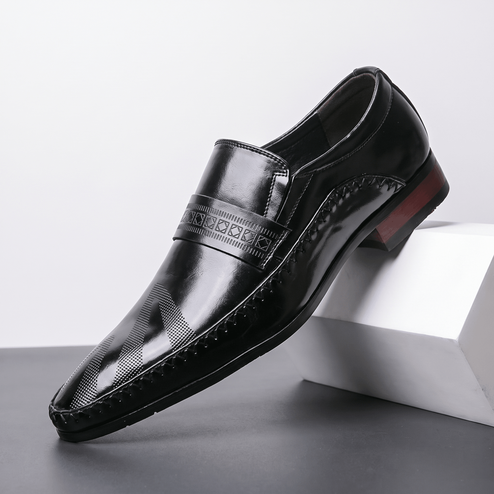 Single black patent leather Leonardo loafer with etched band and burgundy heel accent, propped against a white block in studio setting