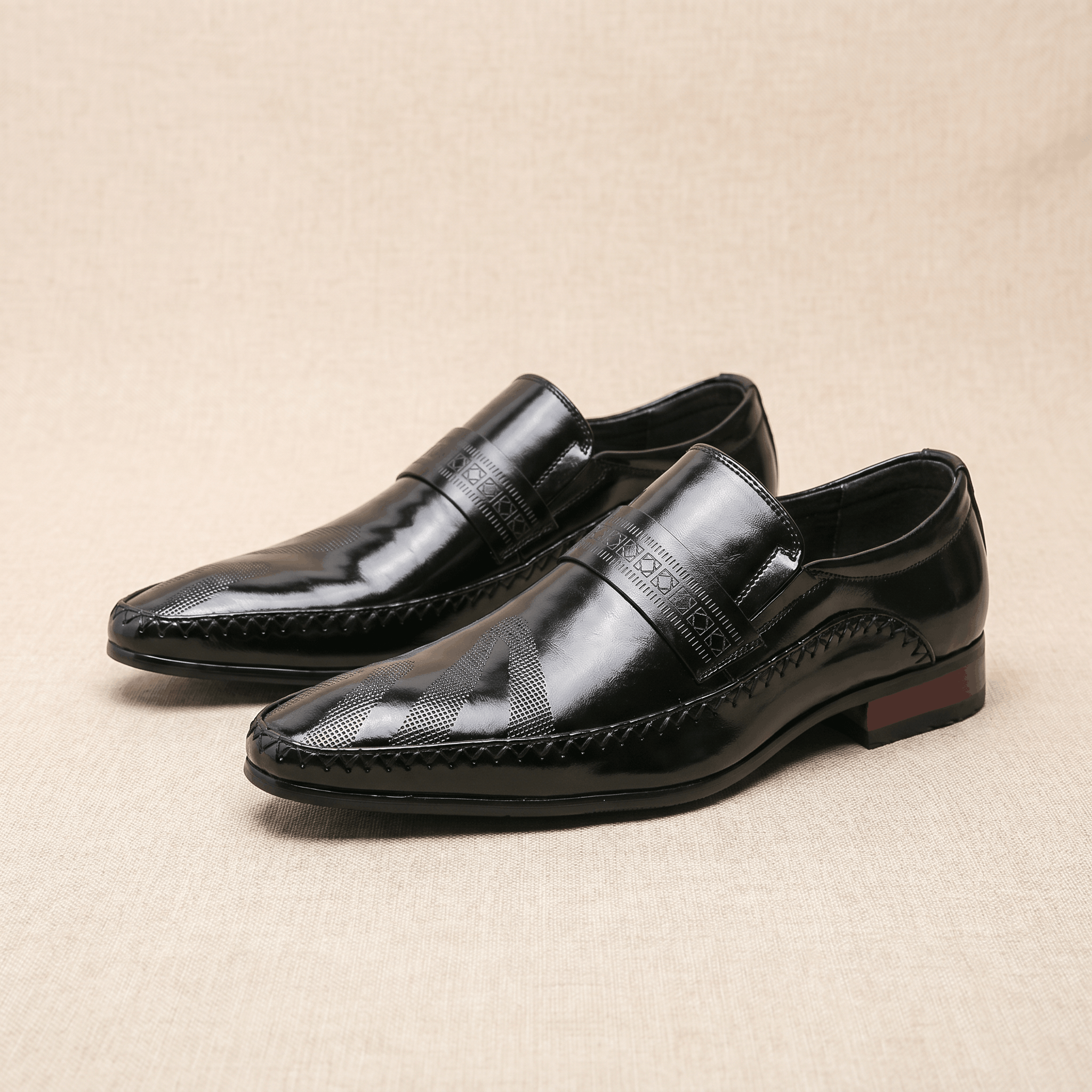 Pair of black patent leather Leonardo loafers with etched band and burgundy heel accent on beige backdrop
