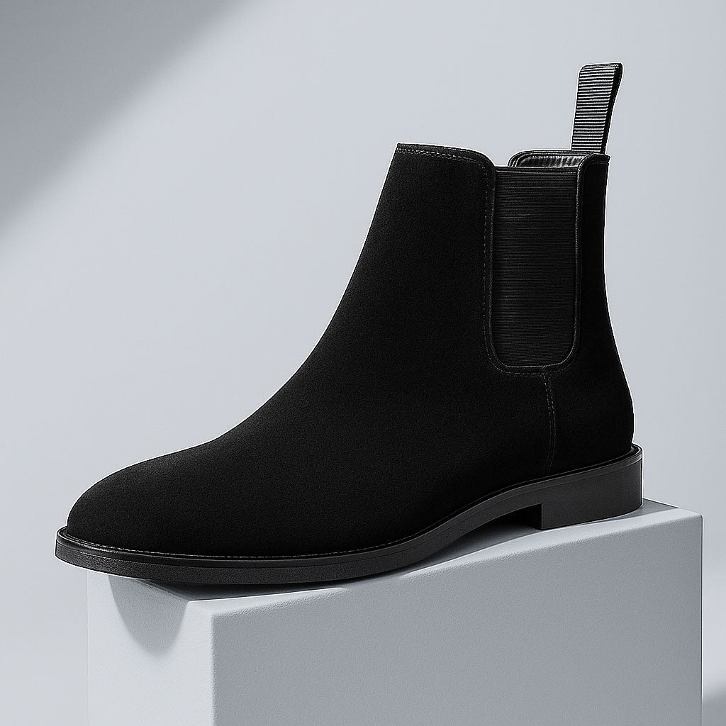 A black suede Chelsea boot resting on a white block against a light gray backdrop.