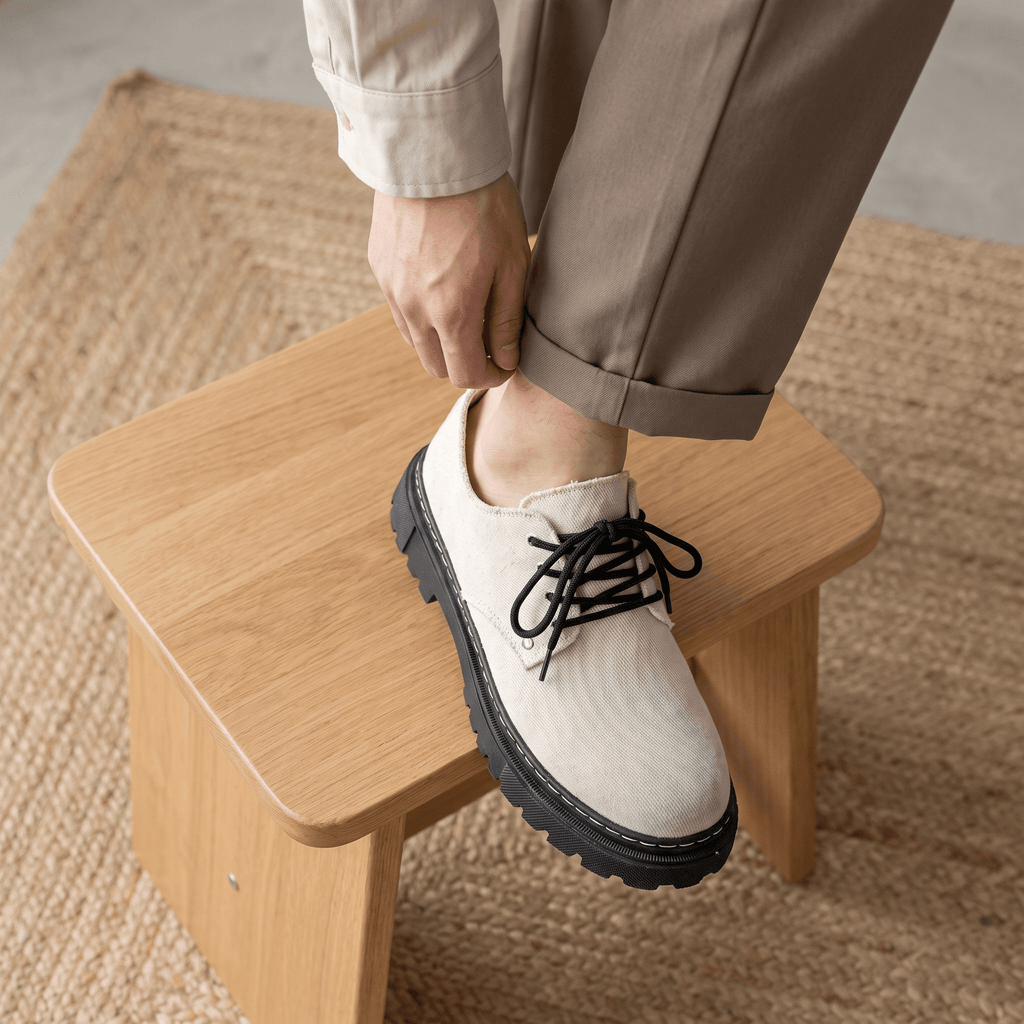 Man adjusting tan trouser cuff with foot resting on wooden stool wearing beige denim Manuel shoe over jute rug