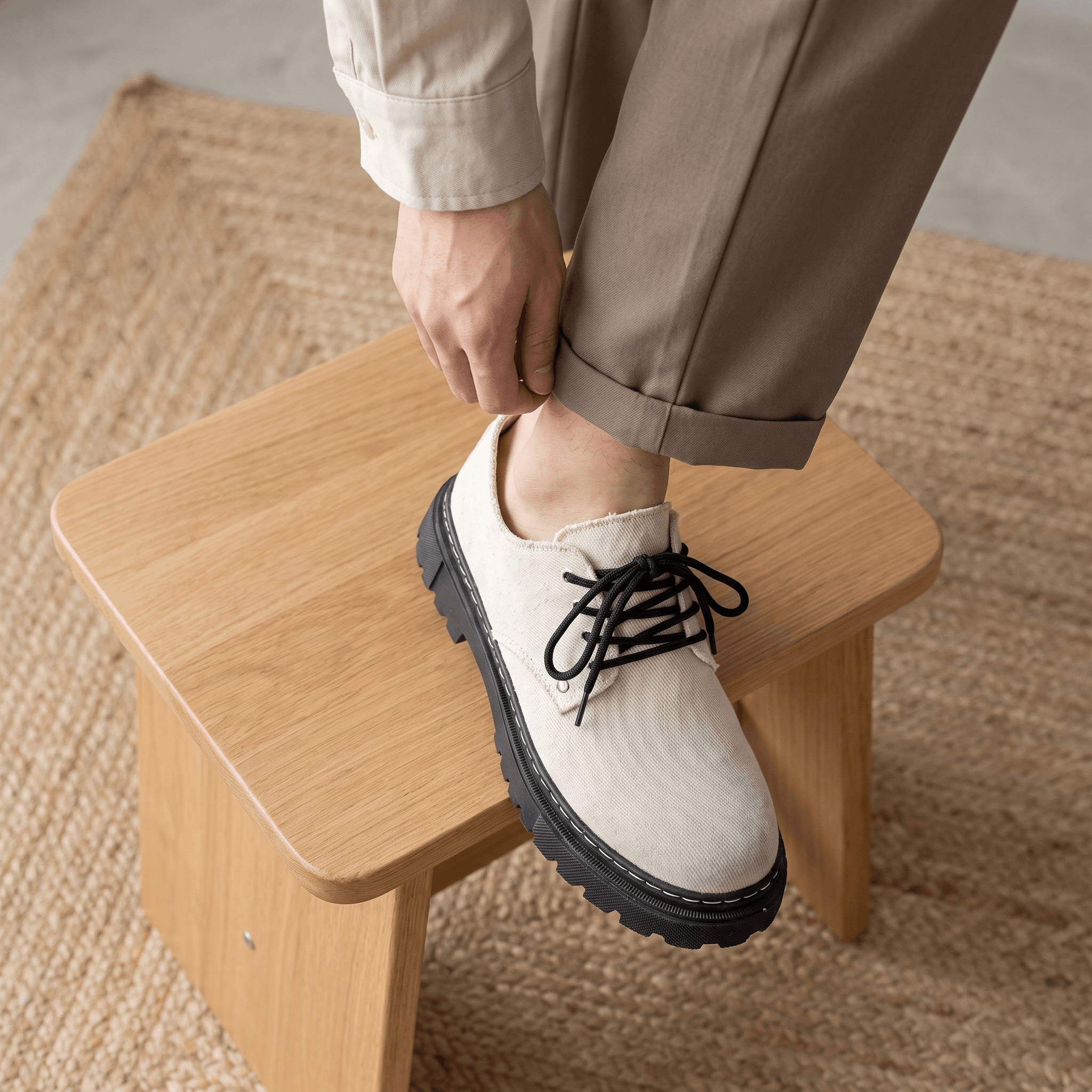 Man adjusting tan trouser cuff with foot resting on wooden stool wearing beige denim Manuel shoe over jute rug