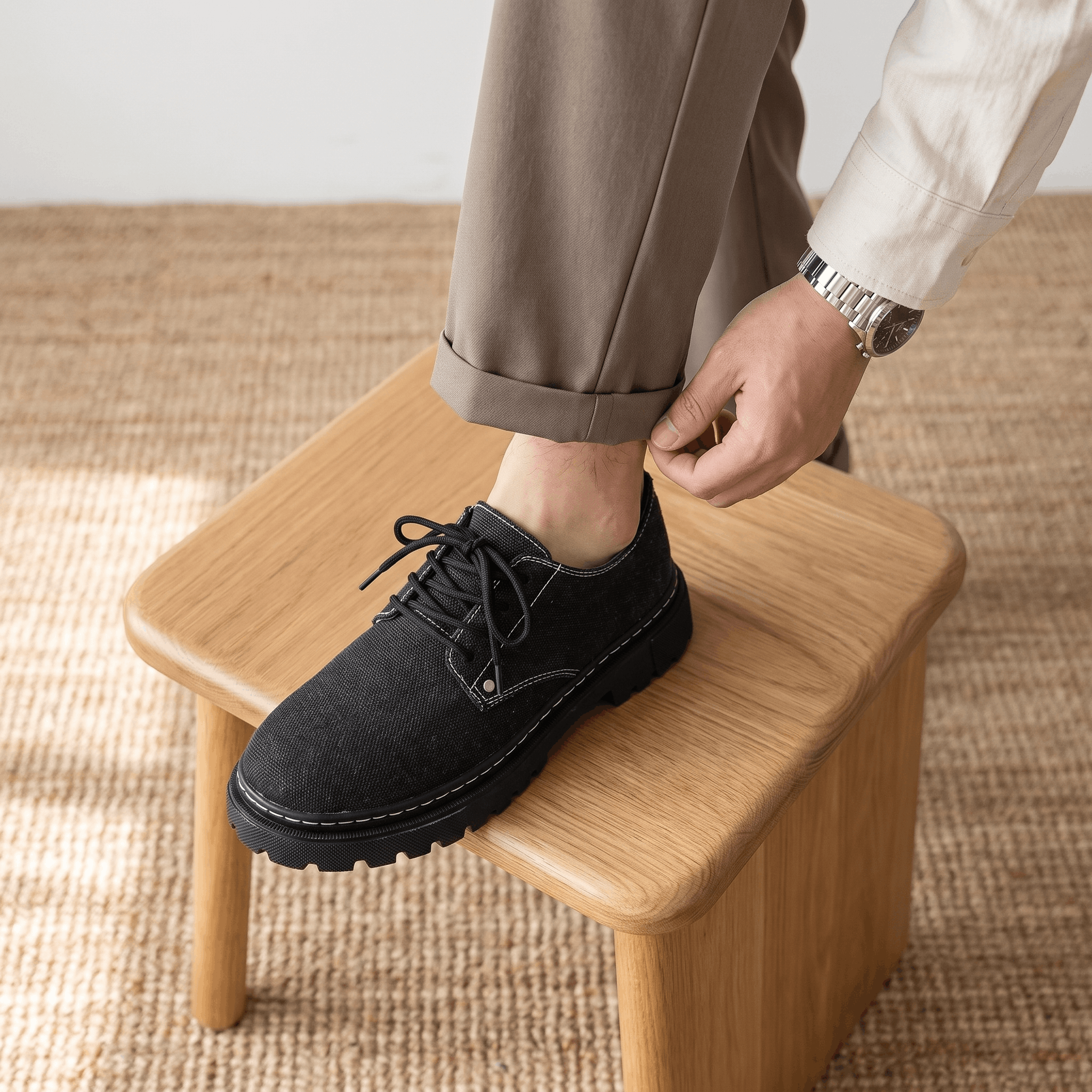 Man adjusting tan trouser cuff with foot resting on wooden stool wearing black denim Manuel shoe over jute rug