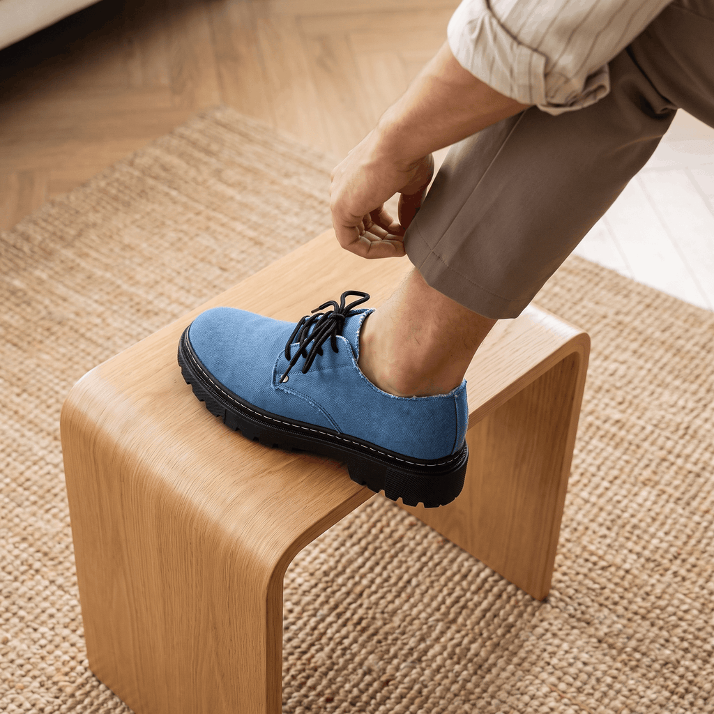 Man tying laces on blue denim Manuel shoe with foot resting on wooden stool over jute rug