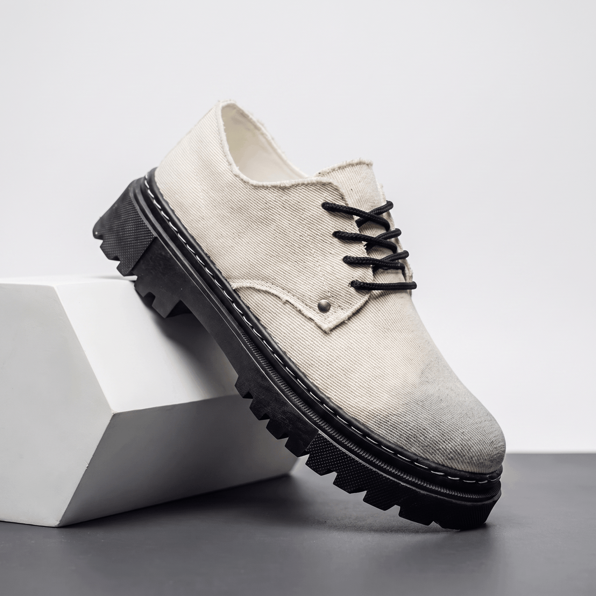 Single beige denim Manuel lace-up shoe with black rubber lug sole, propped against a white block in studio setting