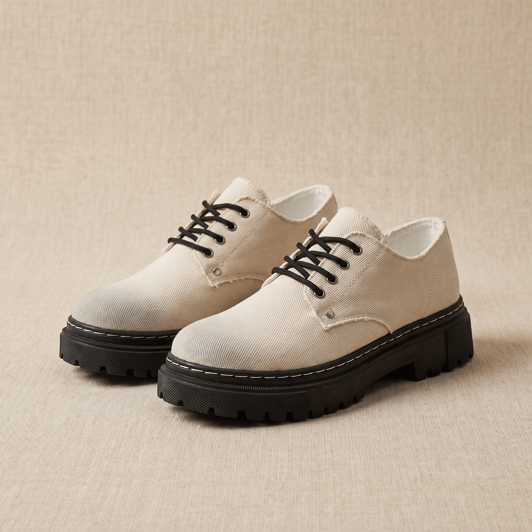 Pair of beige denim Manuel lace-up shoes with black rubber lug soles on beige backdrop