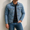 Man wearing a light wash denim jacket with a button-front closure, chest flap pockets, and classic collar over a black T-shirt and blue jeans.