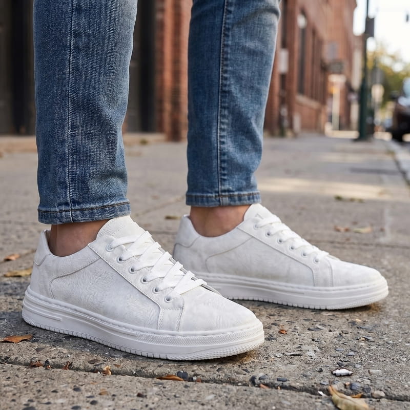Person wearing white leather sneakers with jeans, standing on a paved street with fallen leaves