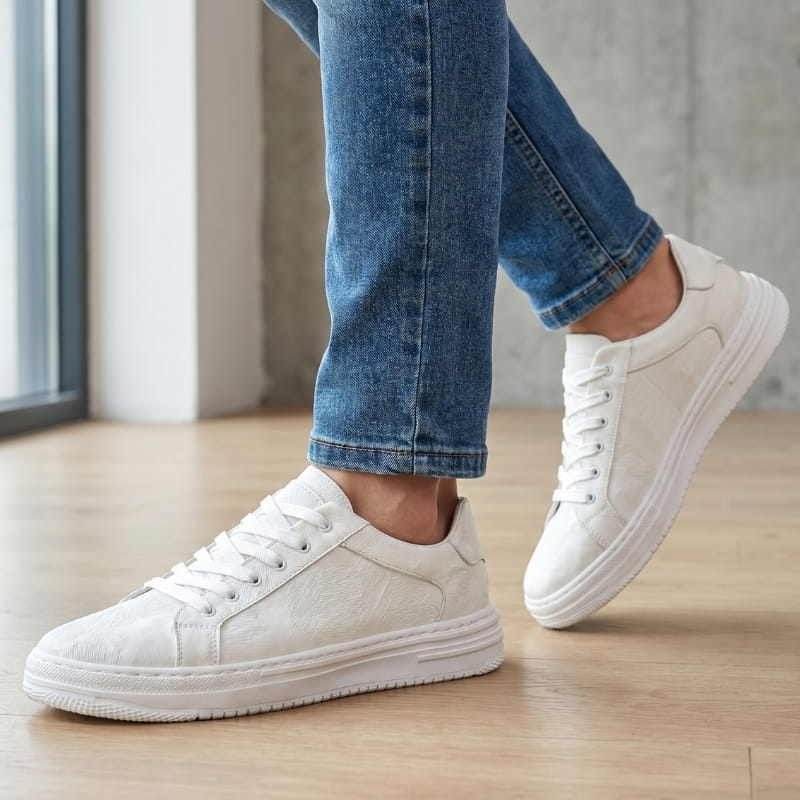 Person wearing white leather sneakers with jeans, walking on a wood floor indoors near a window
