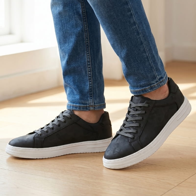 Person wearing black leather sneakers with jeans, standing on a wood floor near a window indoors