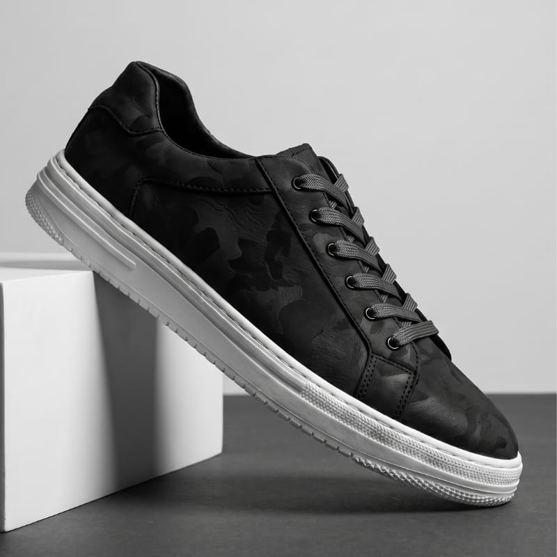 Black leather sneaker with subtle camo texture and white sole propped diagonally against a white block, monochrome studio shot