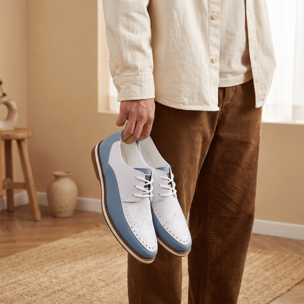 Man in beige shirt holding a pair of white and blue two-tone Mauro brogues by the laces, standing in a sunlit room on a jute rug