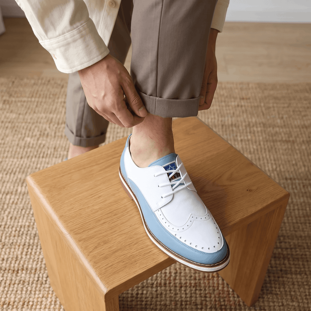 Man adjusting trouser cuff with foot on wooden block, wearing white and blue two-tone Mauro brogue over a jute rug