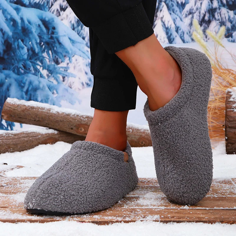 A model's ankles wearing gray teddy fleece slides, standing on a snowy wooden bench outdoors.