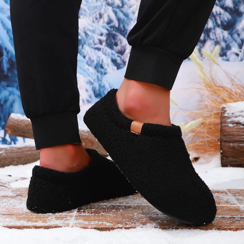 Close-up of a model's ankles wearing black teddy fleece slides, standing on a snowy wooden bench outdoors.