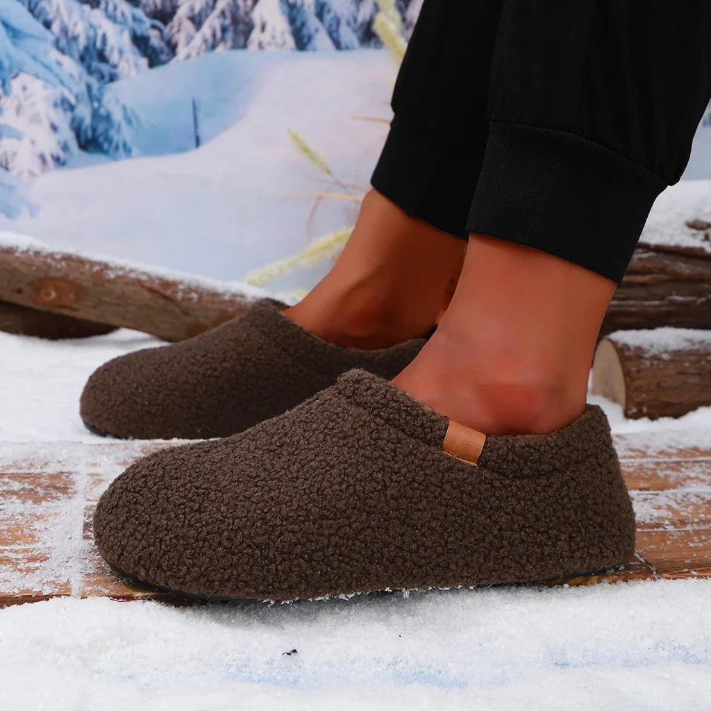 A model's ankles wearing brown teddy fleece slides, standing on a snowy wooden bench outdoors.