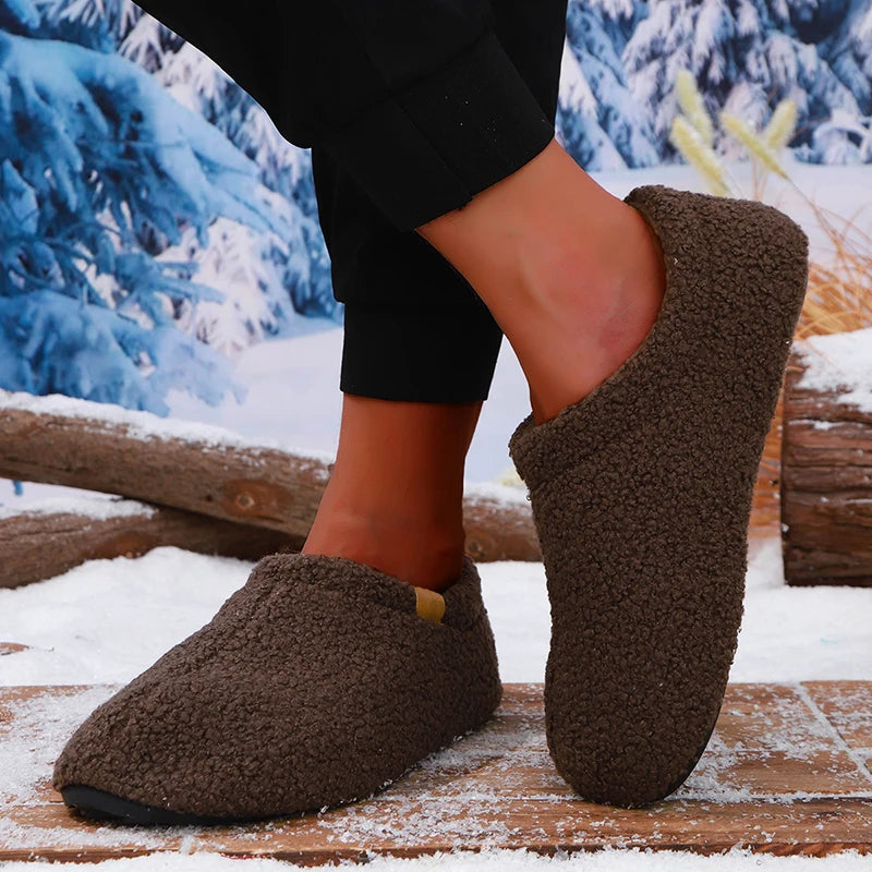Close-up of a model's ankles in brown teddy fleece slides, walking on a snow-dusted wooden bench outdoors.