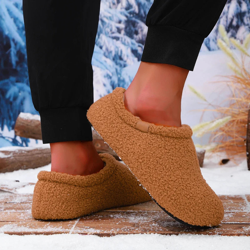 A person mid-step wearing tan teddy fleece slides, one foot lifted, on a snowy wooden bench outdoors.