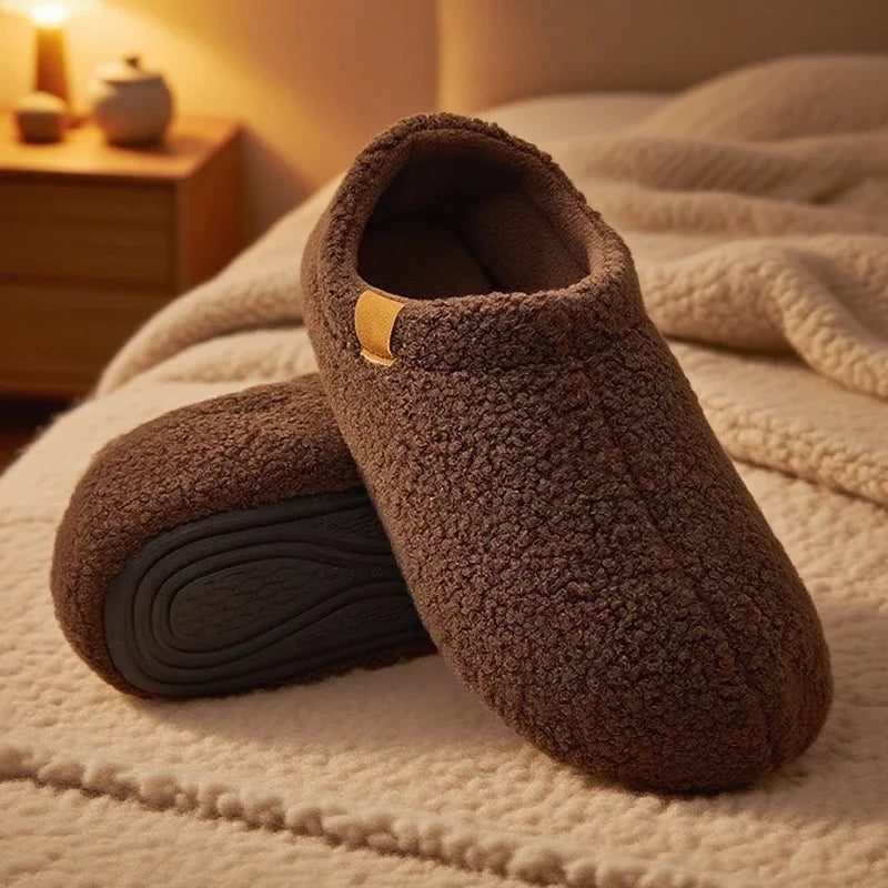 Pair of brown teddy fleece slides with anti-slip soles, resting on a bed with a cozy blanket in warm ambient lighting.