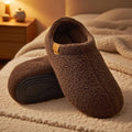 Pair of brown teddy fleece slides with anti-slip soles, resting on a bed with a cozy blanket in warm ambient lighting.
