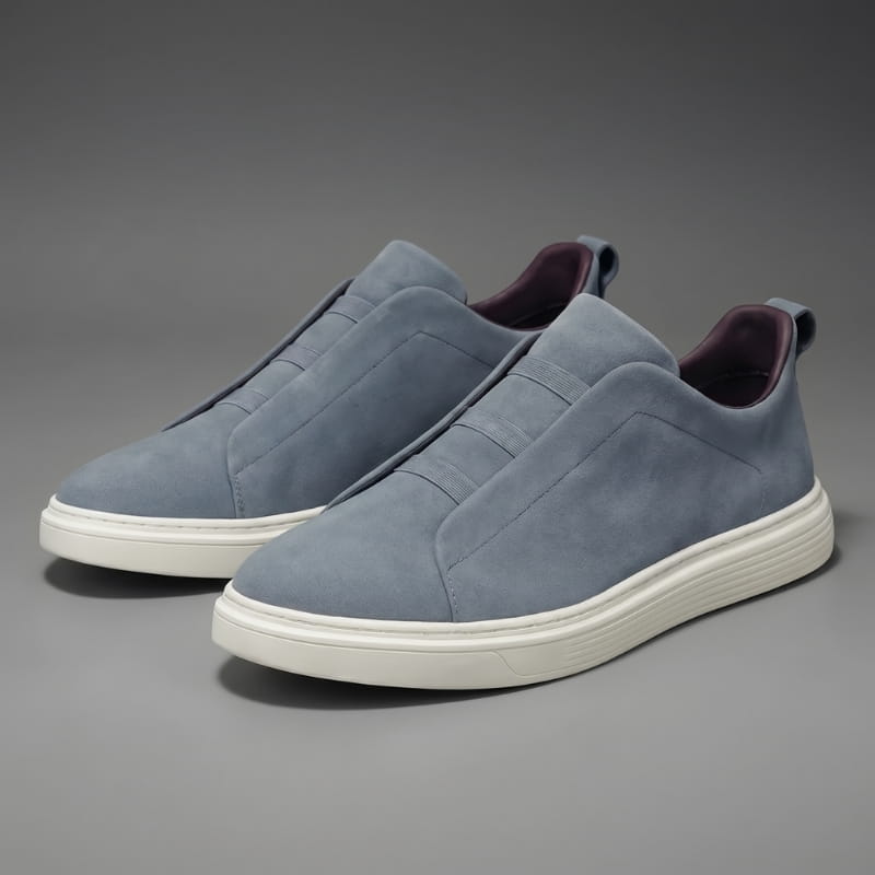 Pair of blue-gray suede slip-on sneakers with white soles facing forward on a gray studio background
